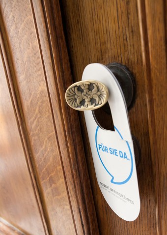 Am Türknauf einer alten Holztür hängt ein weißer Türhänger mit dem Spruch "Für Sie da" in einer Sprechblase.