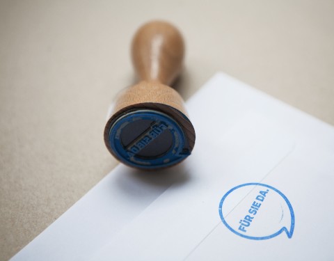 Ein Stempel liegt mit der sichtbaren Stempelseite auf der Seite. Auf einem Blatt ist der Stempelabdruck des Sprechblasen-Logos "Für Sie da" zu sehen.